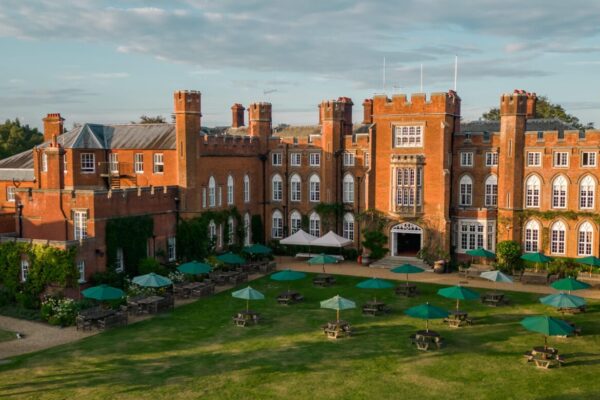 Cumberland Lodge in the sunshine, pictured from the air by a drone. The red brick building is surrounded by grass and trees and on the lawn there are tables with green umbrellas up.