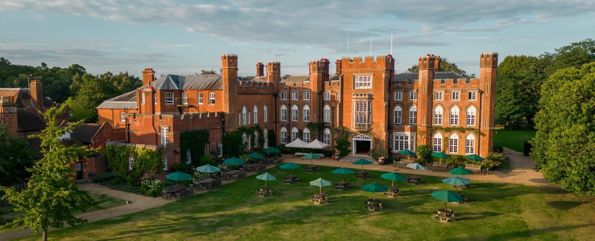 Cumberland Lodge in the sunshine, pictured from the air by a drone. The red brick building is surrounded by grass and trees and on the lawn there are tables with green umbrellas up.