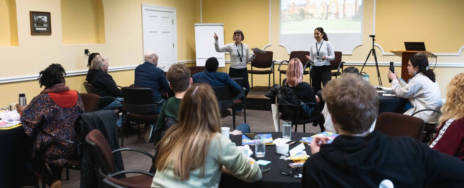 A group session at our Dis/Mis-information workshop. Two members of our Programme team are standing at the front of the room, facing the group of participants who are seated around tables.