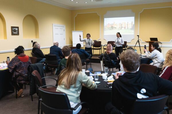 A group session at our Dis/Mis-information workshop. Two members of our Programme team are standing at the front of the room, facing the group of participants who are seated around tables.