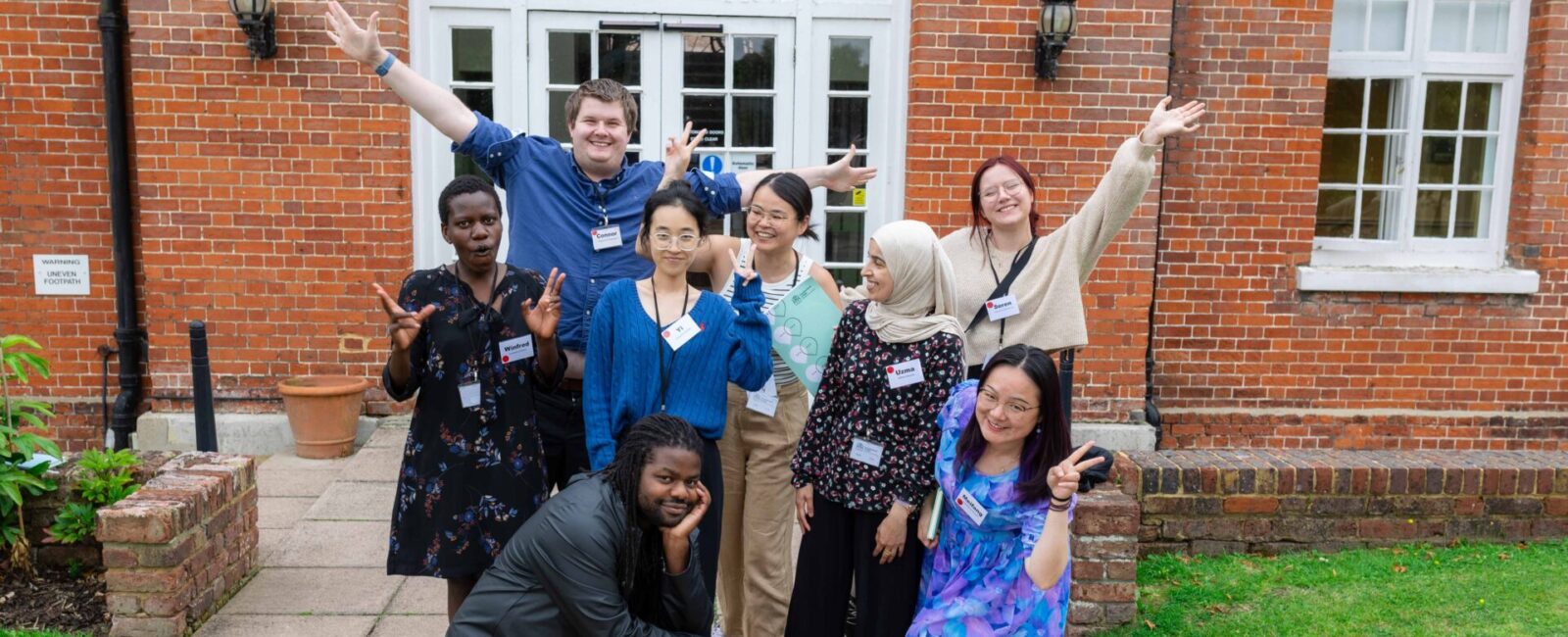 Group of 8 PhD students posing with arms raised on grey path in front of red-bricked building.