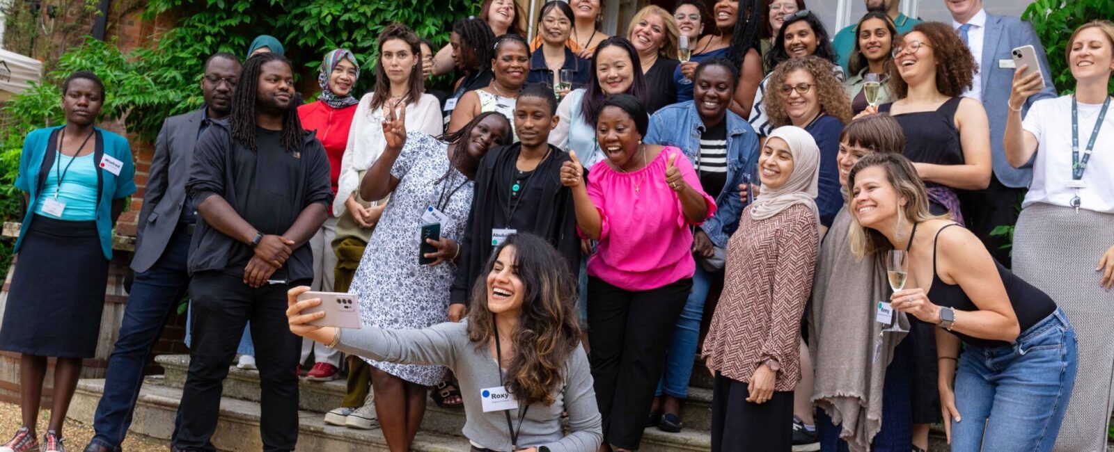 Group of PhD students posing with arms in the air on steps in front of leafy background.