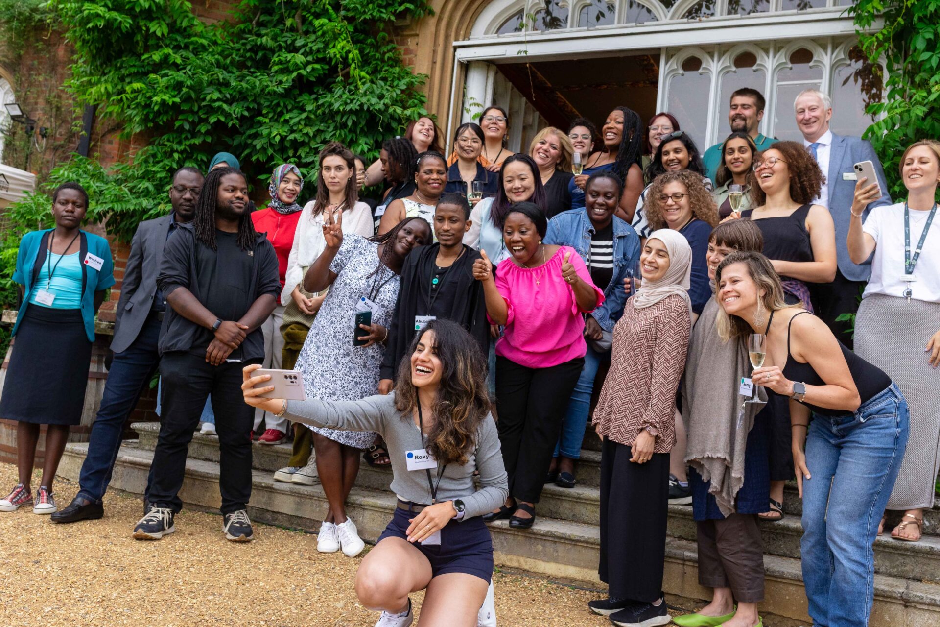 Group of PhD students posing with arms in the air on steps in front of leafy background.