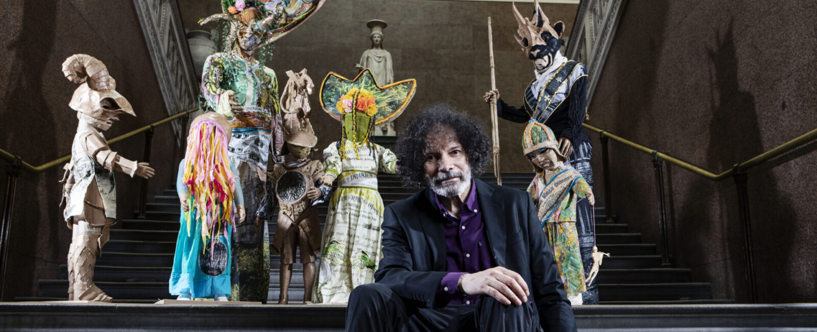 Artist Hew Locke sitting in front of The Watchers, a series of carnivalesque figures installed around the museum galleries.