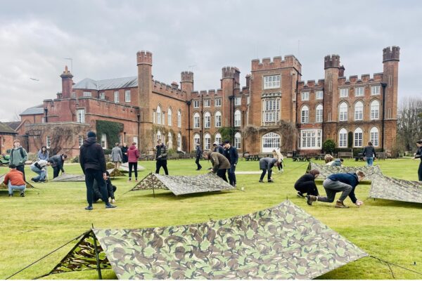 A group taking part in a team-building activity involving tents on the lawn with Cumberland Lodge in the background.
